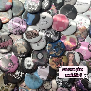 May include: A collection of circular pin-back buttons featuring diverse graphic designs, including anime characters, animals, text like 'i girls', 'Clairo', and 'custom pins available!', and various patterns in a wide array of colors and styles.