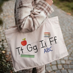 May include: A cream-colored canvas tote bag featuring a graphic print of the letters Tt, Gg, Ii, Ff, and ABC in black and blue, accompanied by a red apple, a stack of three books, and a green ruler on a lined background.
