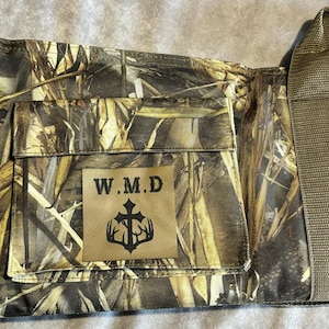 May include: Camouflage pattern bag with a pocket and a handle. The bag features a patch with the letters "W.M.D" and a cross-like emblem. The dominant colors are shades of brown, green, and tan, creating a natural, outdoor aesthetic.