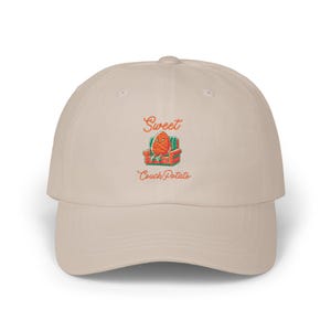 Puede incluir: Gorra de béisbol beige con visera curva. La gorra presenta un gráfico de un personaje de batata sentado en una silla, con las palabras "Sweet Couch Potato" en rojo y verde.