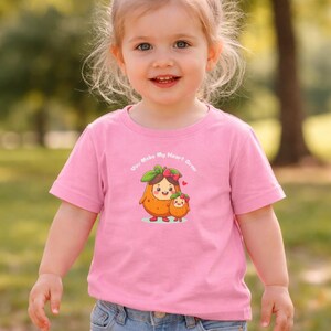 Può includere: Una bambina indossa una maglietta rosa e pantaloncini di jeans, sorridendo alla fotocamera. La maglietta presenta un'illustrazione di due personaggi di frutta con la scritta "You Make My Heart Grow". Un fiocco rosa adorna i capelli della bambina.