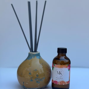May include: A ceramic essential oil diffuser with reeds and a brown glass bottle. The diffuser is a round, yellow vase with blue accents and a floral design. The bottle has a black cap and a label that reads "Ruby Bliss".