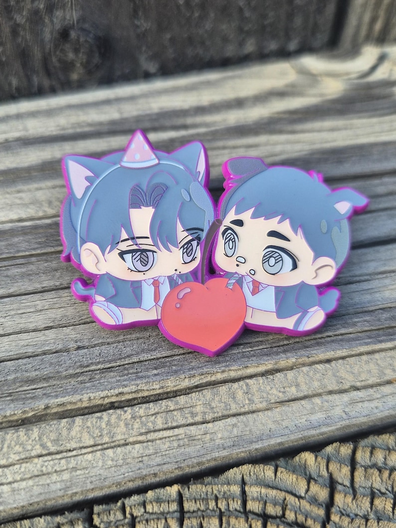Op de afbeelding: Twee stripfiguren met kattenoren en een hartvormig object. De figuren dragen pakken en feesthoeden. Het hart is rood met een roze omtrek. De figuren zijn in tinten blauw, roze en wit.