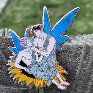 Könnte beinhalten: Ein Anstecknadel mit zwei Figuren in hellblauen, drapierten Gewändern mit blauen Feenflügeln, die auf einer Sonnenblume sitzen. Die Figuren umarmen sich zärtlich. Die Anstecknadel hat eine kupferfarbene Umrandung.