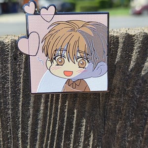 May include: A square enamel pin depicting a cartoon character with brown hair and clothing. The character is smiling, with pink hearts around them. The pin has a silver border and a pink background.
