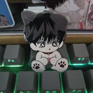 Könnte beinhalten: Ein Emaille-Pin mit einer Cartoonfigur mit schwarzen Haaren und Hundeohren. Die Figur sitzt mit pfotenförmigen Füßen und einem roten Kragen. Der Pin befindet sich auf einer grauen Tastatur mit grüner Hintergrundbeleuchtung.