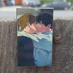 Op de afbeelding: Een rechthoekige pin met een illustratie van twee kussende mensen. E&eacute;n persoon heeft bruin haar, de ander zwart. Het kunstwerk is in mangastijl, met zachte schaduwen en een focus op de gezichten.