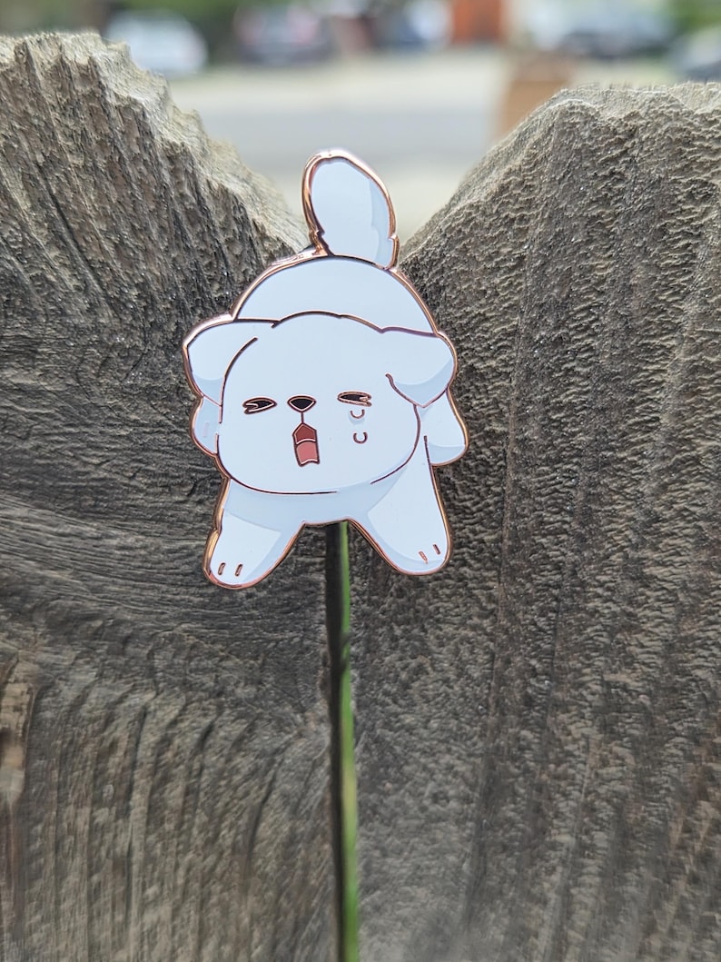Op de afbeelding: Een lichtblauwe emaille pin in de vorm van een cartoonhond met een verdrietige uitdrukking. De pin heeft een ros&eacute;gouden omtrek en een pin back. De hond huilt en heeft zijn mond open. De pin is bevestigd aan een groene stengel.