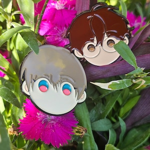 Puede incluir: Dos pines de esmalte con caras de dibujos animados de personas con cabello marrón y gris. Los pines están colocados sobre un fondo de flores rosas y follaje verde.