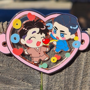 May include: A heart-shaped pin with a pink border and a clear center. Inside, two cartoon figures are depicted, one in brown and the other in blue, with hearts and other colorful shapes. The pin is attached to a green stem.