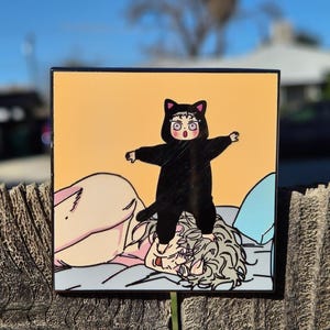 Op de afbeelding: Vierkante kunstdruk met een cartoonillustratie van een persoon in een zwart kattenkostuum die op een ander persoon in bed staat. De achtergrond is lichtoranje.