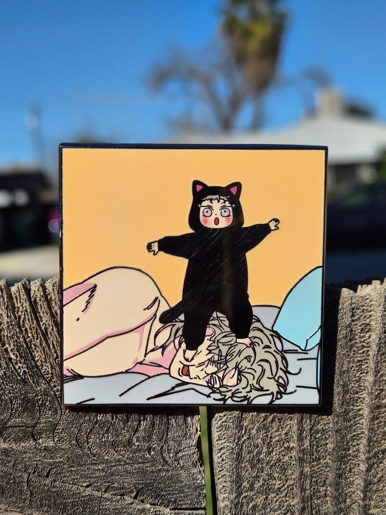 Op de afbeelding: Vierkante kunstdruk met een cartoonillustratie van een persoon in een zwart kattenkostuum die op een ander persoon in bed staat. De achtergrond is lichtoranje.