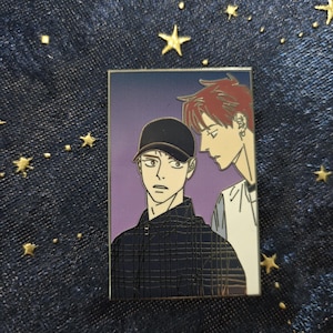 Op de afbeelding: Een gouden emaille pin met twee mannen. De man links draagt een zwart-wit geruit overhemd en een zwarte baseballpet. De man rechts draagt een wit overhemd en heeft rood haar. De achtergrond is een paarse gradiënt.