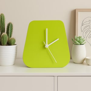 May include: A lime green, modern-style clock with white hands sits on a white surface. A cactus in a white pot, a framed leaf print, a small succulent, and a candle are also on the surface.