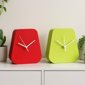May include: Two modern, square-shaped clocks, one red and one lime green, with white hands. The clocks are on a white surface, with potted plants in the background. The clocks are a stylish home decor item.