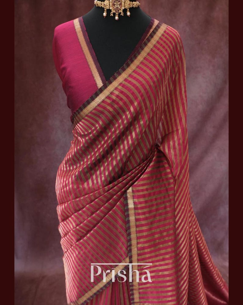 May include: A red and gold striped saree with a matching blouse and a gold necklace. The saree features vertical stripes and is draped on a mannequin. The word "Prisha" is visible on the saree.