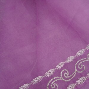 Pu&ograve; includere: Un tessuto trasparente color lavanda con un delicato bordo ricamato. Il bordo presenta un motivo ripetuto di disegni floreali e a volute bianche. La parola "Blouse" e il marchio "Prisha" sono stampati sul tessuto.