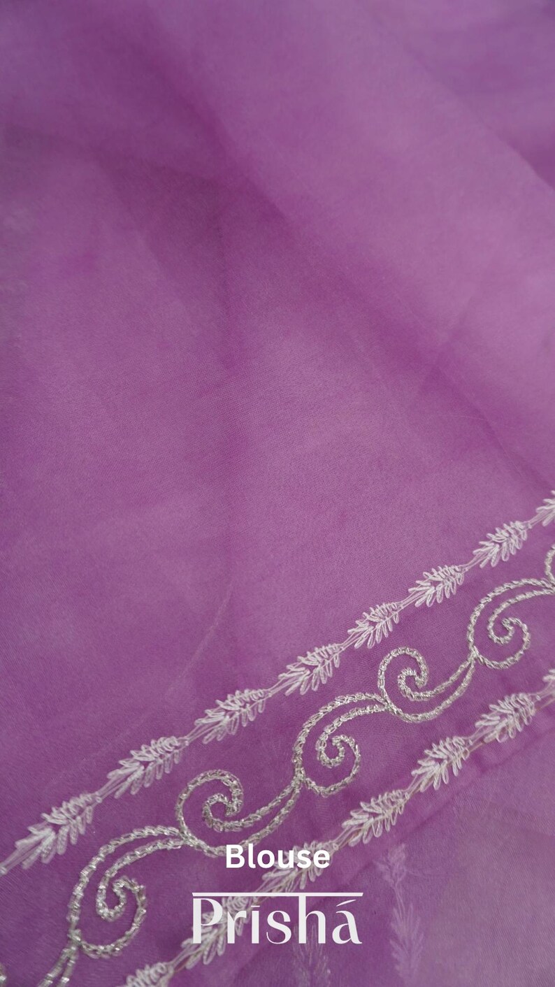 Pu&ograve; includere: Un tessuto trasparente color lavanda con un delicato bordo ricamato. Il bordo presenta un motivo ripetuto di disegni floreali e a volute bianche. La parola "Blouse" e il marchio "Prisha" sono stampati sul tessuto.