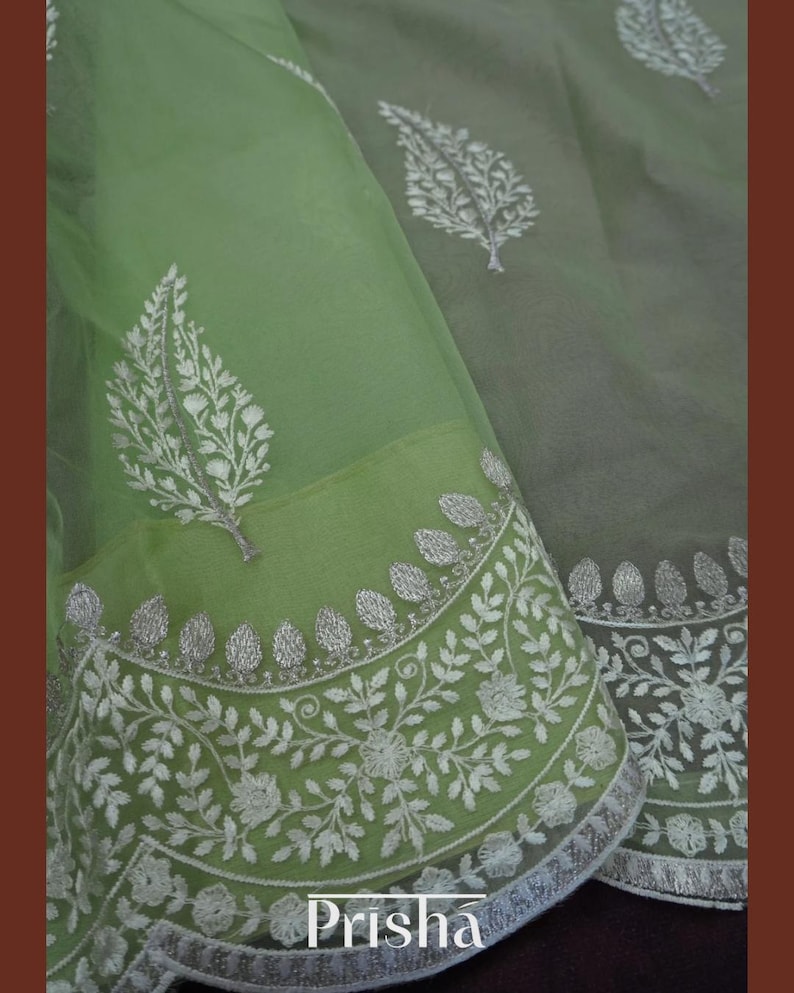 Pu&ograve; includere: Primo piano di un tessuto verde chiaro trasparente con intricati ricami floreali bianchi. Il tessuto presenta un bordo dettagliato con motivi floreali e di foglie. La parola "Prisha" &egrave; visibile nella parte inferiore dell'immagine.