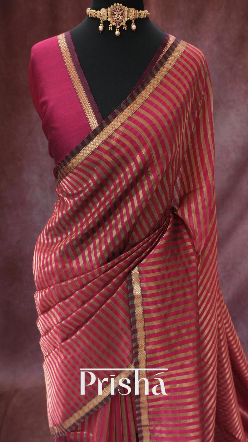 May include: A draped magenta saree with gold stripes and trim, displayed on a mannequin. The saree features the brand name "Prisha" at the bottom. A matching magenta blouse and ornate gold necklace complete the ensemble.