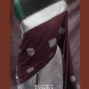 May include: A maroon silk saree with a silver border and intricate silver designs. The saree is draped on a mannequin and has a green blouse.