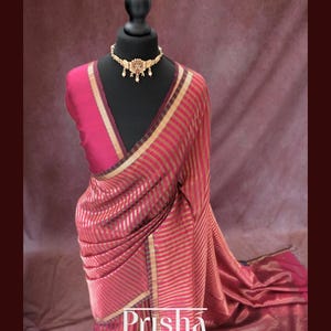 May include: A red and gold striped saree draped on a black mannequin. The saree features a gold border and is paired with a matching blouse. A gold necklace with intricate detailing adorns the mannequin's neck. The word "Prisha" is displayed on the saree.