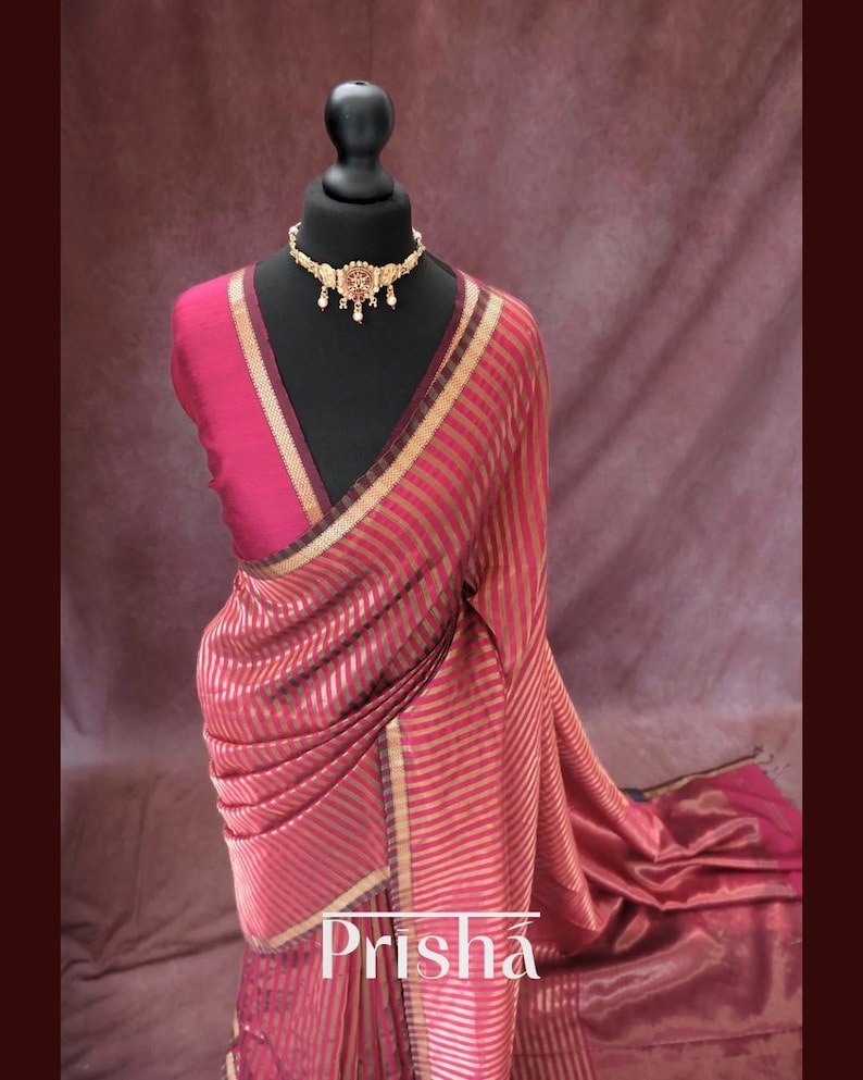 May include: A red and gold striped saree draped on a black mannequin. The saree features a gold border and is paired with a matching blouse. A gold necklace with intricate detailing adorns the mannequin's neck. The word "Prisha" is displayed on the saree.