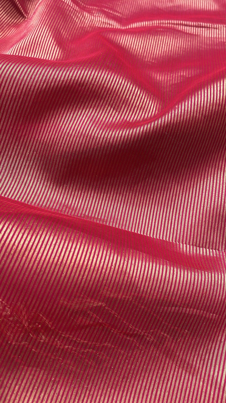 May include: Close-up of a vibrant pink fabric with a subtle sheen. The material features a pattern of thin, vertical, metallic gold stripes, creating a textured and luxurious appearance. The fabric is likely used for apparel or home decor.