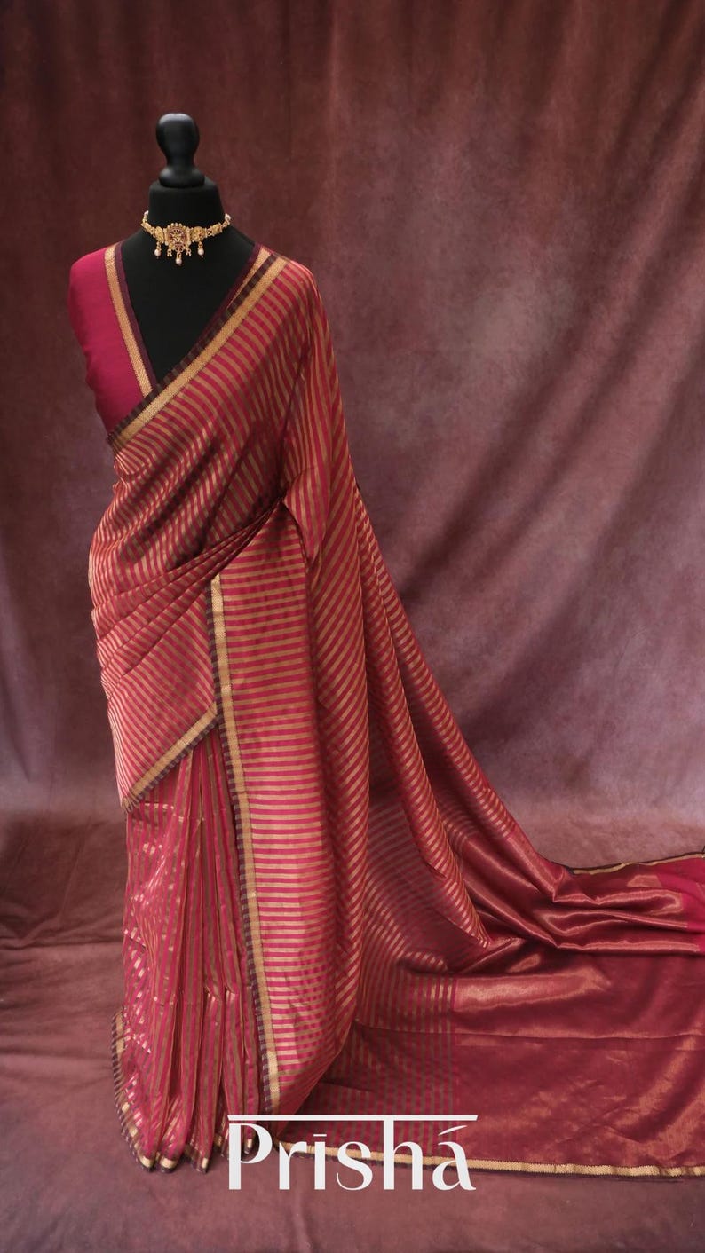 May include: A red and gold striped saree draped on a mannequin. The saree has gold trim and a matching blouse. A gold necklace adorns the mannequin's neck. The word "Prisha" is visible at the bottom of the image.