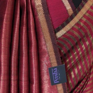 May include: Close-up of a maroon saree with gold-colored trim and vertical stripes. The fabric has a subtle sheen and a textured appearance. The brand name "Prisha" is visible on a tag.