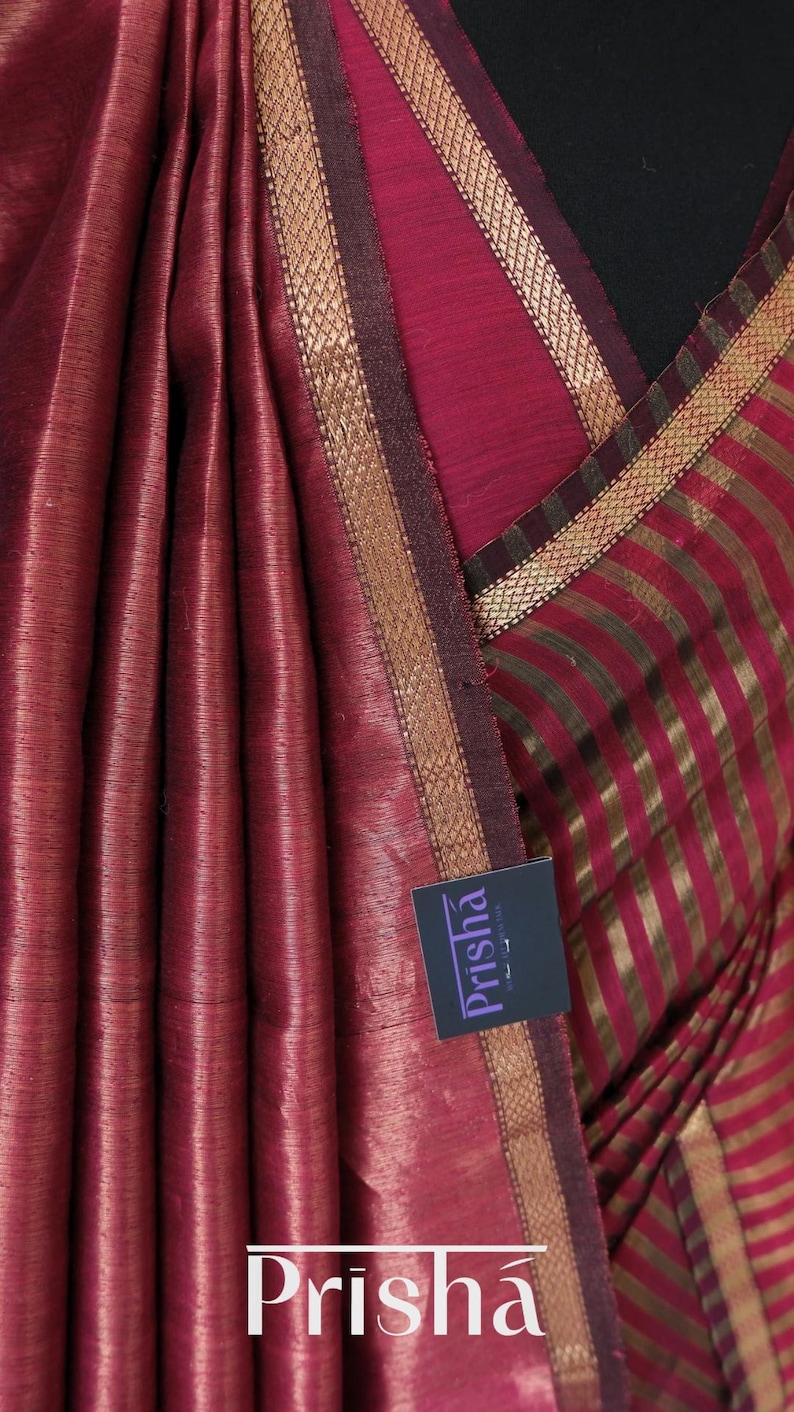 May include: Close-up of a maroon saree with gold-colored trim and vertical stripes. The fabric has a subtle sheen and a textured appearance. The brand name "Prisha" is visible on a tag.