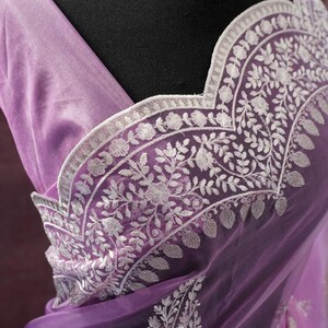 Pu&ograve; includere: Un sari lavanda con ricami floreali bianchi intricati. Il sari presenta un tessuto delicato e trasparente e un bordo smerlato. La parola "Prisha" &egrave; stampata in basso a destra del sari.