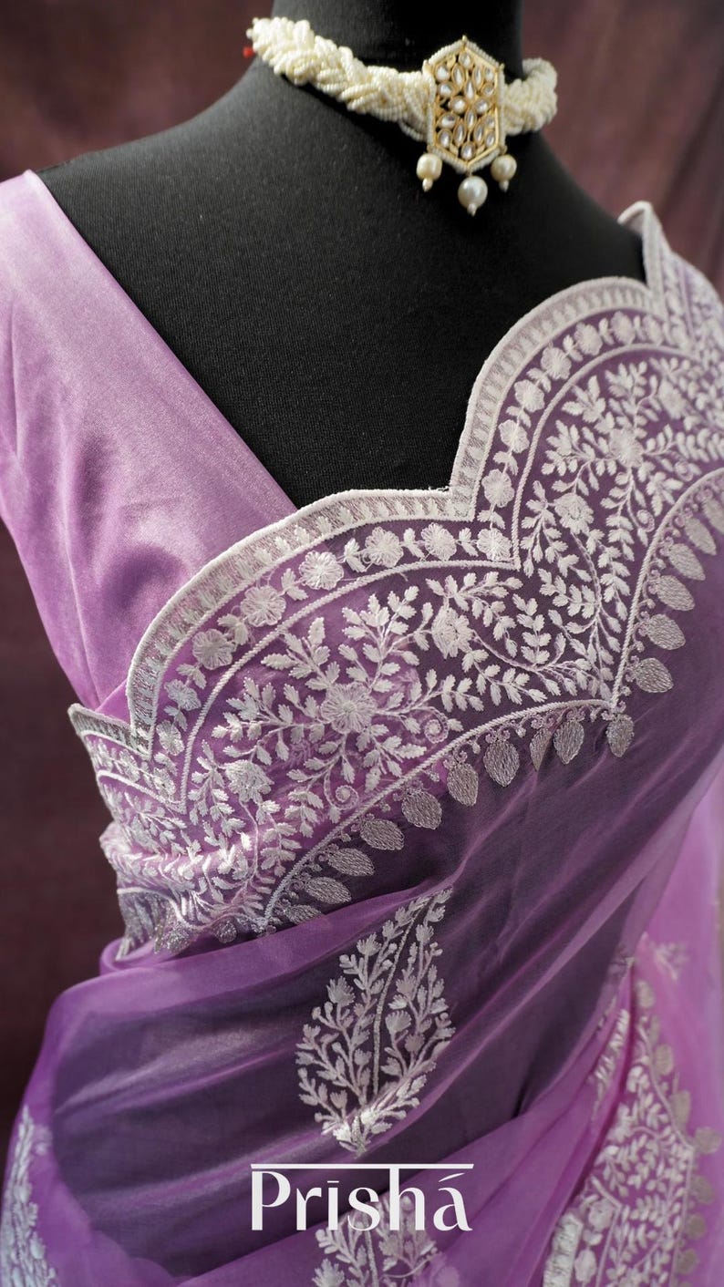 Pu&ograve; includere: Un sari lavanda con ricami floreali bianchi intricati. Il sari presenta un tessuto delicato e trasparente e un bordo smerlato. La parola "Prisha" &egrave; stampata in basso a destra del sari.