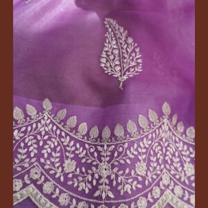 Pu&ograve; includere: Primo piano di un tessuto trasparente color lavanda con ricami bianchi intricati. Il design presenta motivi floreali e a foglie lungo il bordo e sparsi sulla superficie. La parola "Prisha" &egrave; visibile in basso al centro.