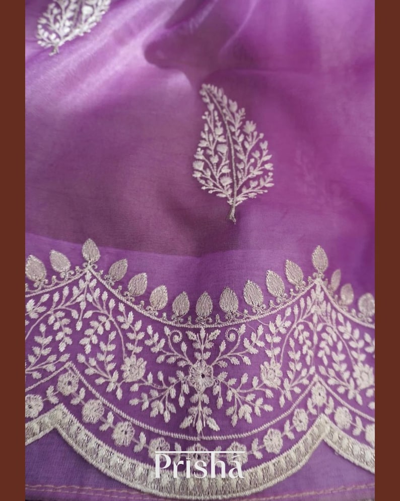 Pu&ograve; includere: Primo piano di un tessuto trasparente color lavanda con ricami bianchi intricati. Il design presenta motivi floreali e a foglie lungo il bordo e sparsi sulla superficie. La parola "Prisha" &egrave; visibile in basso al centro.