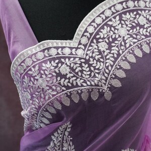 Pu&ograve; includere: Un sari color lavanda con ricami floreali bianchi intricati. Il sari presenta un tessuto delicato e trasparente ed &egrave; adornato con motivi dettagliati lungo i bordi e lo scollo. &Egrave; visibile anche una collana di perline bianche con un ciondolo.