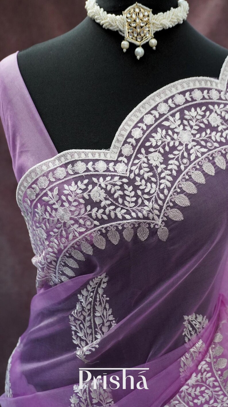 Pu&ograve; includere: Un sari color lavanda con ricami floreali bianchi intricati. Il sari presenta un tessuto delicato e trasparente ed &egrave; adornato con motivi dettagliati lungo i bordi e lo scollo. &Egrave; visibile anche una collana di perline bianche con un ciondolo.