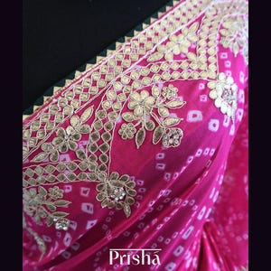 May include: A vibrant pink saree with intricate gold embroidery and floral patterns. The saree features a white and pink tie-dye design and a green border. The word "Prisha" is displayed in white script.