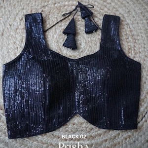 Puede incluir: Una blusa negra sin mangas de lentejuelas con cuello redondo y un lazo de borla decorativo. La blusa tiene el texto "BLACK 02 Prisha" impreso en la parte inferior.