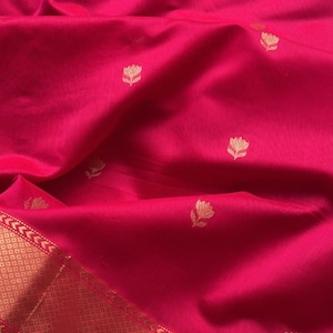 May include: A vibrant fuchsia fabric with a subtle sheen. The textile features a repeating pattern of small, gold-colored floral motifs. A gold border with geometric and arrow designs accents the edge of the fabric.