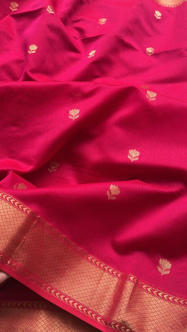 May include: A vibrant fuchsia fabric with a subtle sheen. The textile features a repeating pattern of small, gold-colored floral motifs. A gold border with geometric and arrow designs accents the edge of the fabric.