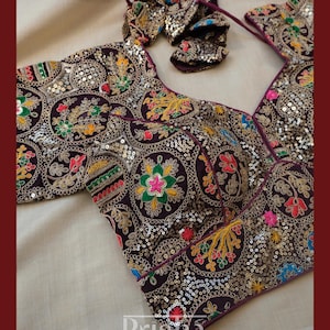 May include: A dark purple blouse featuring intricate circular embroidery with multi-colored floral patterns and silver sequins. The garment has a V-neckline, short sleeves, and a tie-back closure. The word Prisha is printed in white at the bottom.
