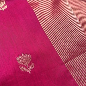 May include: Close-up of a vibrant pink textile with a gold striped border. The fabric features a repeating floral pattern in gold. The textile is likely a silk or similar material, with a subtle sheen.