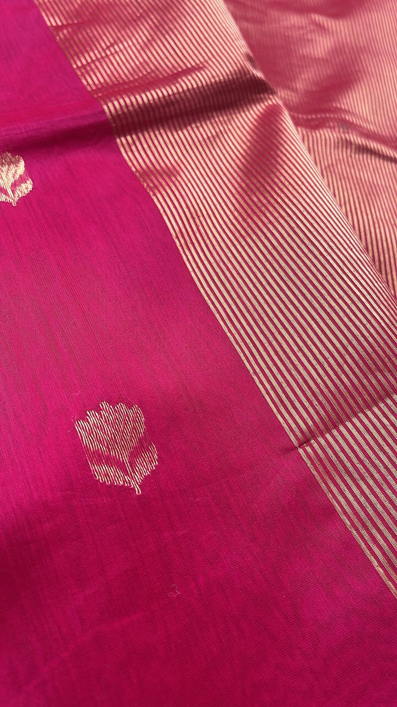 May include: Close-up of a vibrant pink textile with a gold striped border. The fabric features a repeating floral pattern in gold. The textile is likely a silk or similar material, with a subtle sheen.