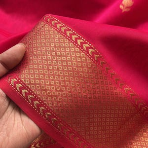May include: Close-up of a vibrant red fabric with a gold patterned border. The border features geometric designs and arrow-like shapes. The fabric has a smooth texture and a subtle sheen.