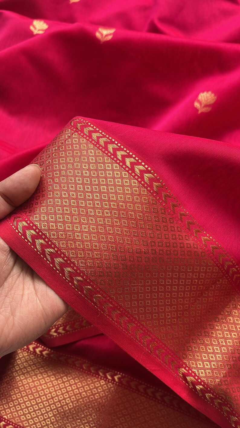 May include: Close-up of a vibrant red fabric with a gold patterned border. The border features geometric designs and arrow-like shapes. The fabric has a smooth texture and a subtle sheen.