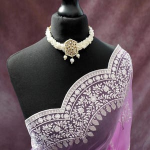 Pu&ograve; includere: Un sari color lavanda con intricati ricami floreali bianchi e una collana di perline bianche abbinata con un ciondolo dorato. Il sari &egrave; drappeggiato su un manichino nero. La parola "Prisha" &egrave; visibile sul sari.