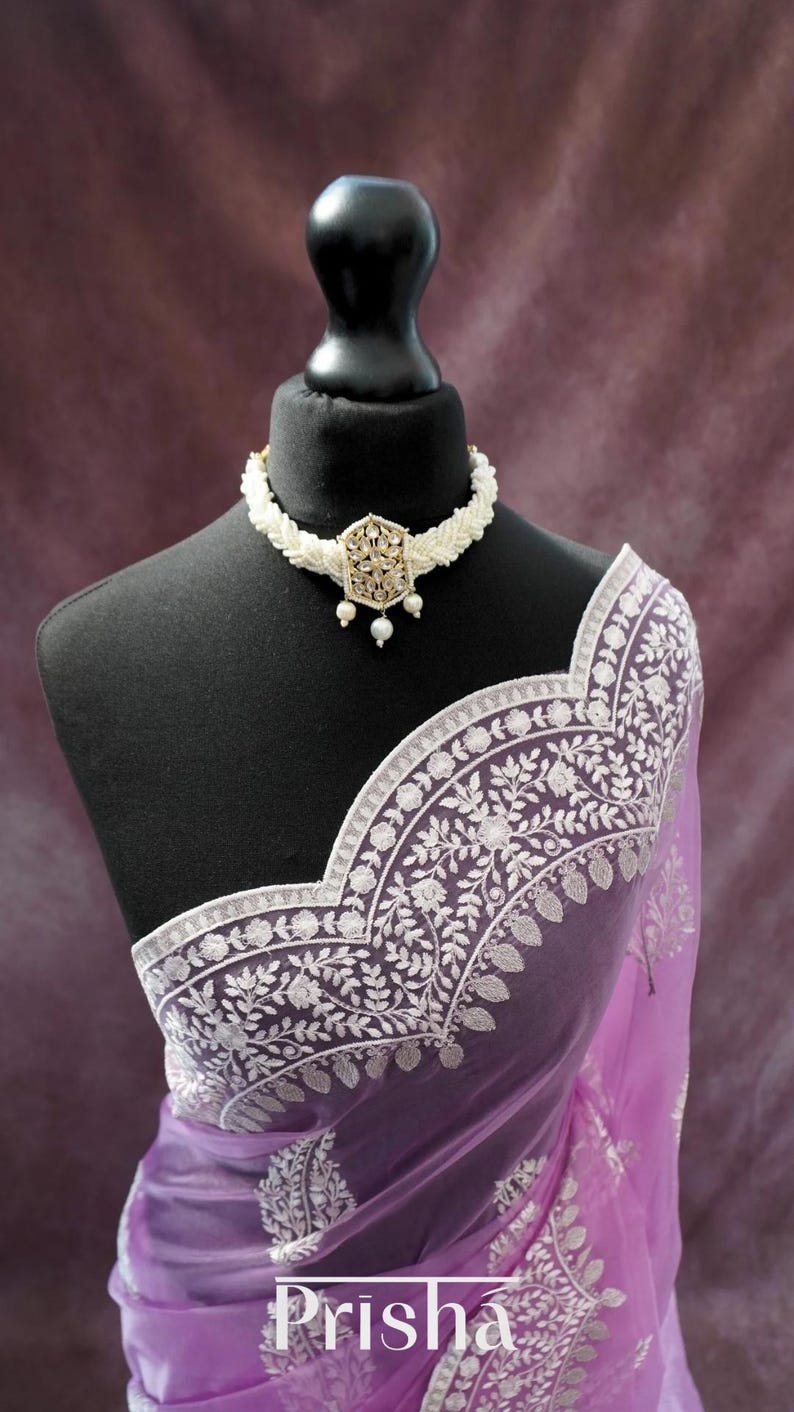 Pu&ograve; includere: Un sari color lavanda con intricati ricami floreali bianchi e una collana di perline bianche abbinata con un ciondolo dorato. Il sari &egrave; drappeggiato su un manichino nero. La parola "Prisha" &egrave; visibile sul sari.