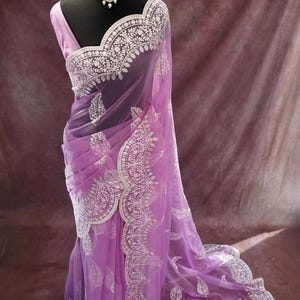 Pu&ograve; includere: Un sari lavanda con ricami bianchi intricati e un bordo smerlato &egrave; drappeggiato su un manichino. Il sari presenta un delicato design floreale e paisley. Una collana di perle con un pendente centrale adorna il collo del manichino. La parola "Prisha" &egrave; visibile in basso.