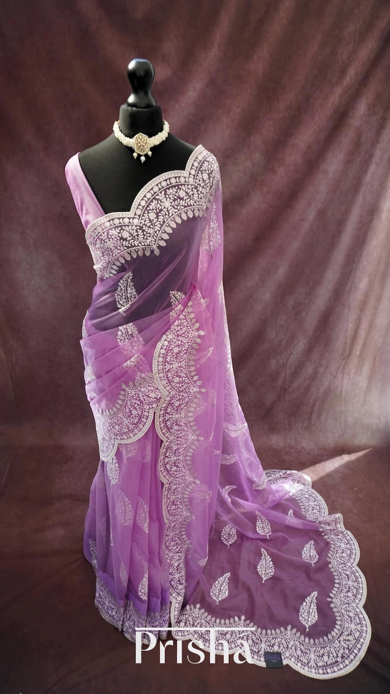 Pu&ograve; includere: Un sari lavanda con ricami bianchi intricati e un bordo smerlato &egrave; drappeggiato su un manichino. Il sari presenta un delicato design floreale e paisley. Una collana di perle con un pendente centrale adorna il collo del manichino. La parola "Prisha" &egrave; visibile in basso.